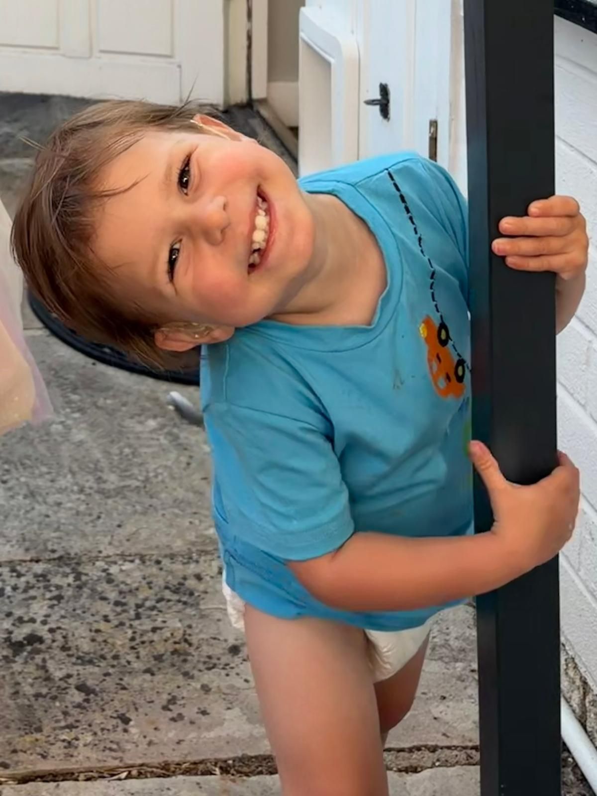 Sam grinning, head tilted, holding onto a doorframe outside the house.
