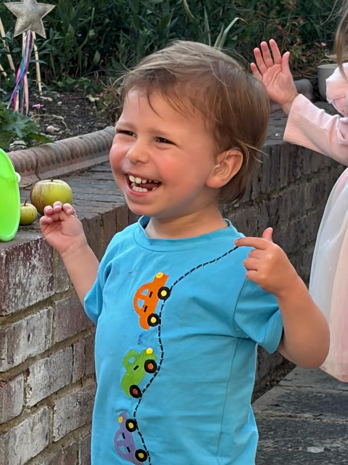 Sam, aged two, laughing in the garden in a bright blue T-shirt printed with cars.